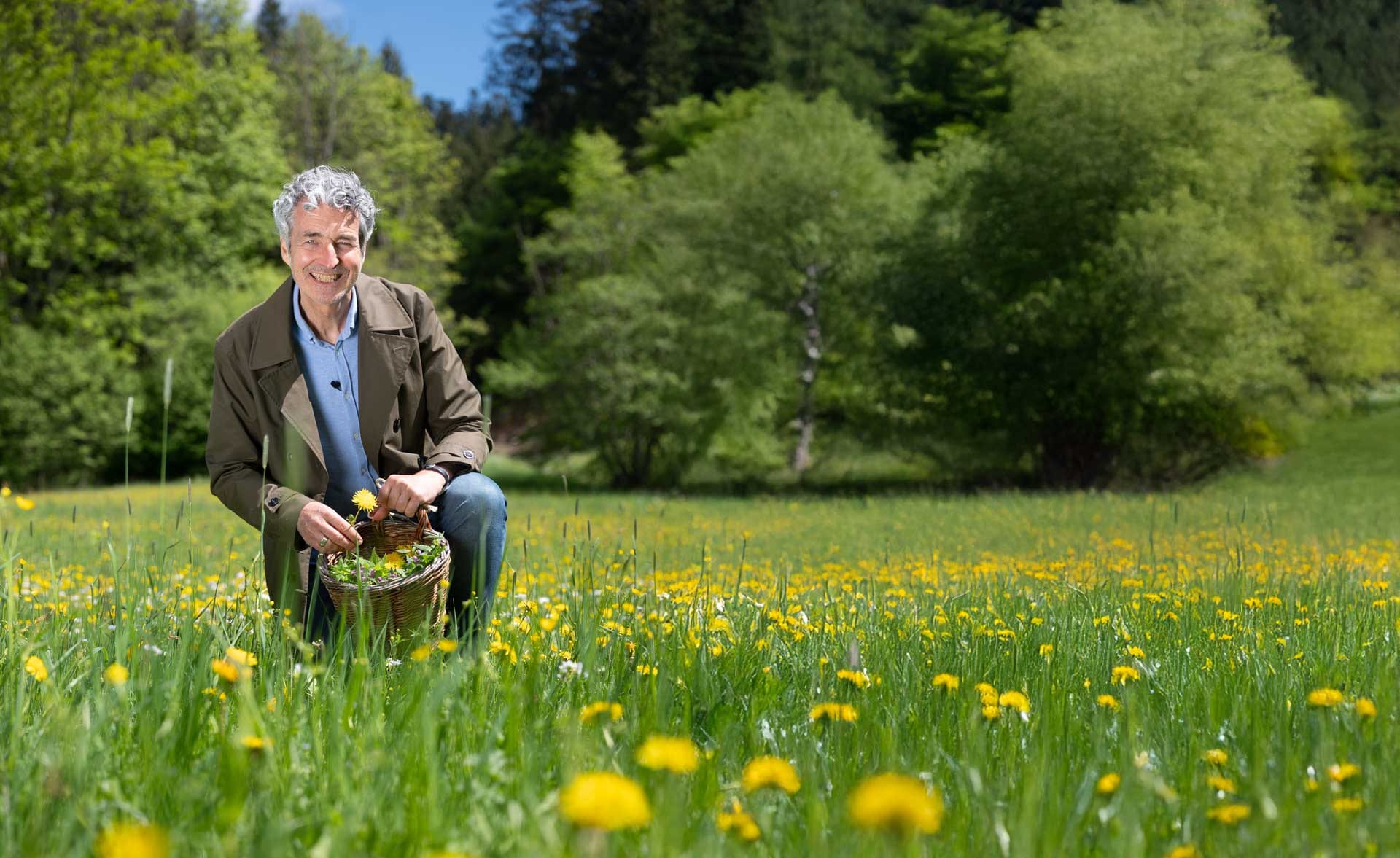 Dr. Markus Strauß kniet lächelnd in einer sonnigen Löwenzahn-Wiese und hält frisch gesammelte Wildpflanzen